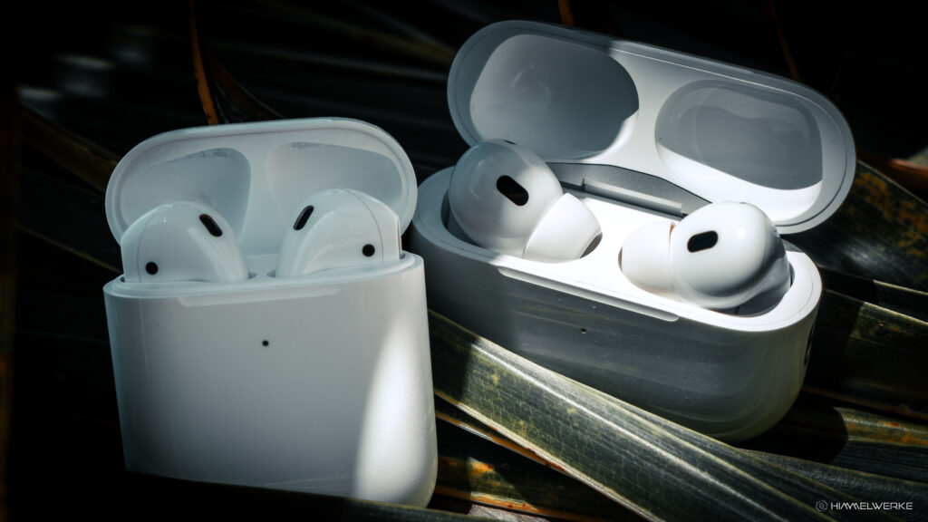 Das Foto zeigt die zwei Ladecase der Apple AirPods der 2. Generation nebeneinander. Links befindet sich die normale Version und rechts die Pro Version. Beide Ladecase sind nach oben hin geöffnet, sodass man die AirPods darin sieht. Beide Case liegen auf einem dunkelgrünem Palmblatt.