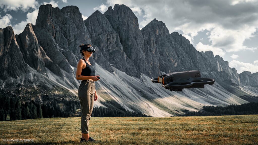 Eine junge Frau steht auf einer Wiese vor einem eindrucksvollen Bergmassiv mit dramatischen Wolkenhimmel. Die Frau hat die DJI FPV Goggle 3 aufgesetzt und in ihrer rechten Hand den DJI Motion RC 3. In der rechten Bildhälfte steht die DJI Avata 2 in der Luft im Vordergrund.