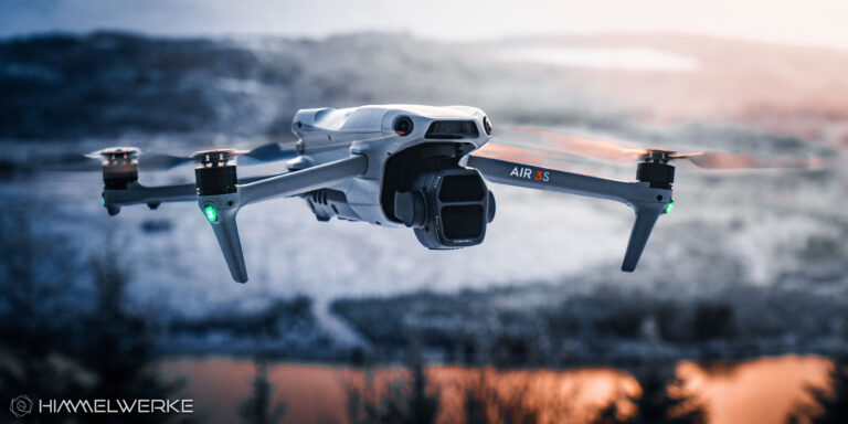 Die DJI Air 3S in der Luft von schräg vorne. Die LED der Drohne leuchten grün. Im Hintergrund ist eine schneebedeckte Hügellandschaft zu erkennen. Im unteren Teil des Bildes leuchtet ein Gewässer orange durch die tief stehende Sonne.