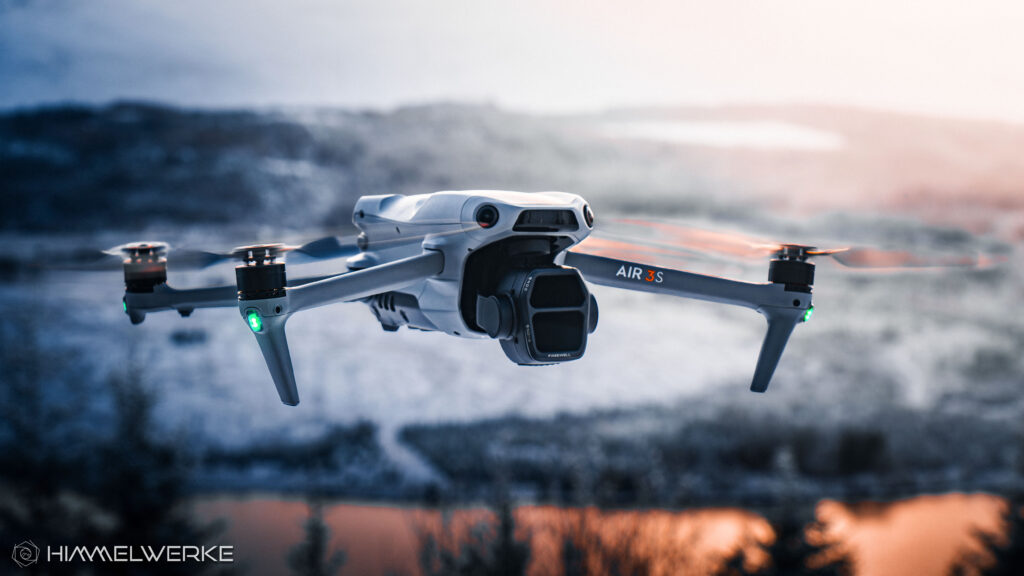 Die DJI Air 3S in der Luft von schräg vorne. Die LED der Drohne leuchten grün. Im Hintergrund ist eine schneebedeckte Hügellandschaft zu erkennen. Im unteren Teil des Bildes leuchtet ein Gewässer orange durch die tief stehende Sonne.