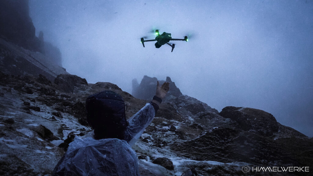 Im Bild ist Martin von Himmelwerke mit einem Regenponcho und einer Kapuze von der rechten Seite zu sehen. Seine linke Hand ist ausgestreckt um nach der am Himmel fliegenden DJI Air 3 S zu greifen, welche sich gerade am Landen befindet. Im Bild kann man eine karge Felslandschaft im Hintergrund erkennen und das Wetter ist sichtlich schlecht. Es liegt etwas Schnee am Boden und es sieht so aus, als ob es schneit/regnet. Die LED der Drohne leuchten hellgrün. Die allg. Lichtstimmung ist düster.