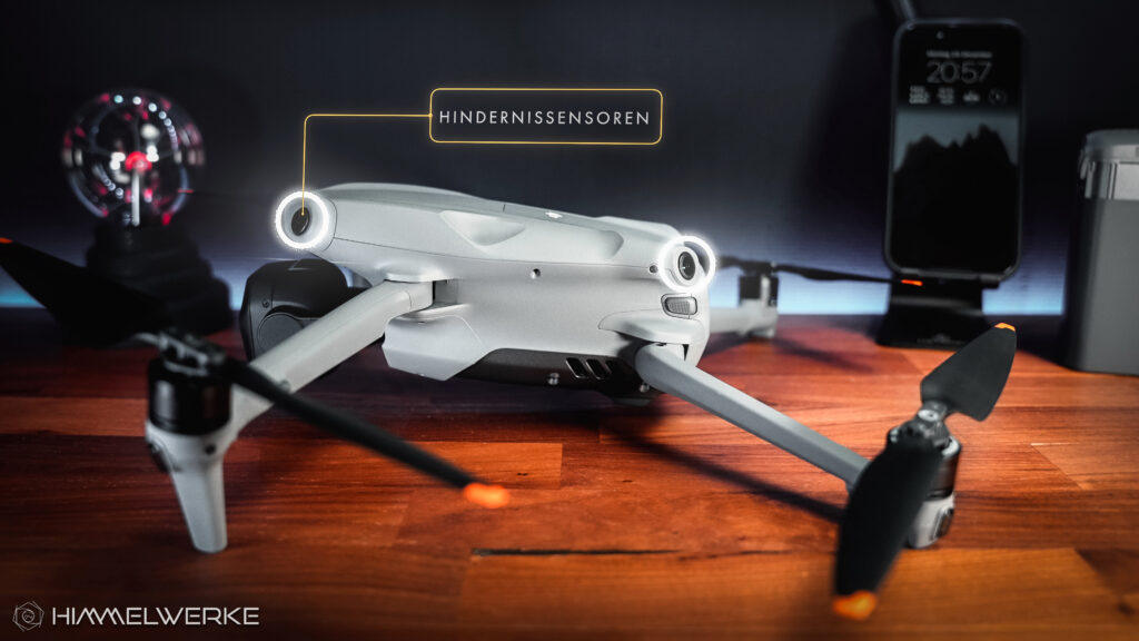 Die DJI Air 3S auf einem Holztisch, ausgeklappt, von der linken Seite. Ein Schriftzug mit "Hindernissensoren" weist mit gelber Umrandung und Linie auf die Hindernissensoren der Drohne hin, welche zusätzlich mit kleinen weiß leuchtenden Kreisen umrandet sind.