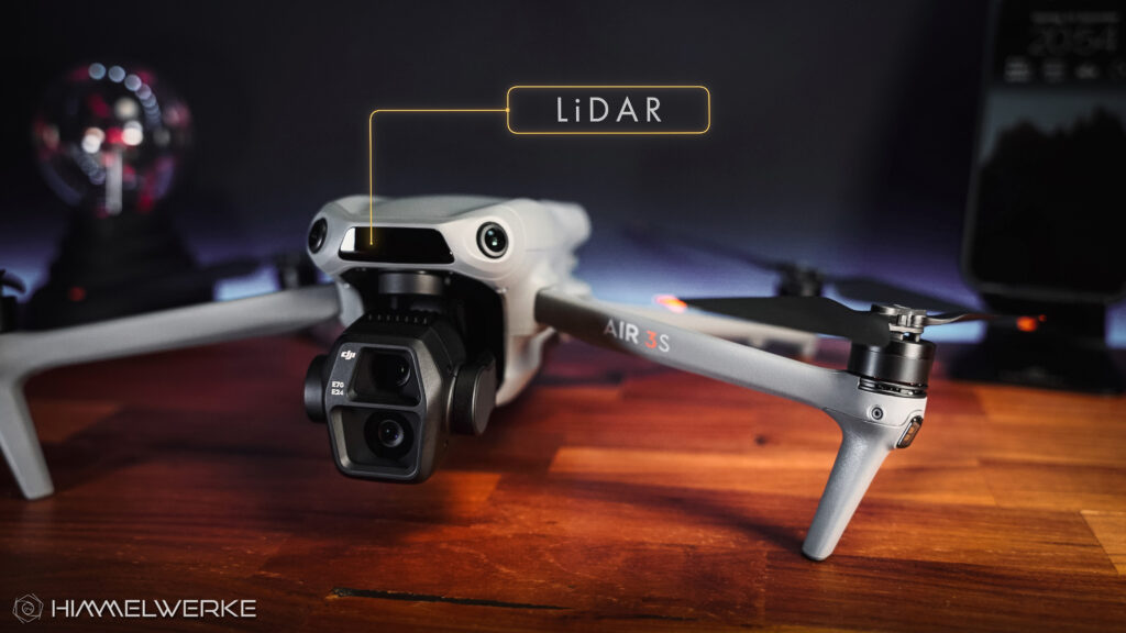 Die DJI Air 3 S ausgeklappt auf einem braunen Holztisch von vorne. Ein Schriftzug mit "LiDAR" weist in einer gelben Umrandung und Linie auf den neuen LiDAR Sensor in der Front der Drohne hin.