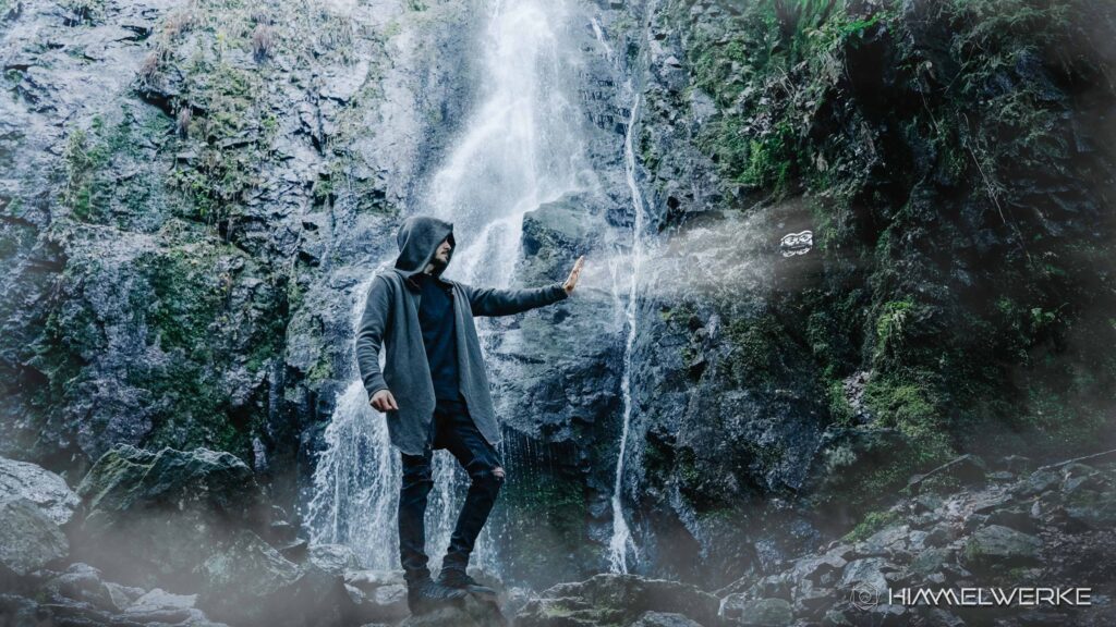 Ein Himmelwerke Bild zum DJI NEO 2 Test wo ein Mann mit Kaputze vor einem mystischen Wasserfall auf einem Stein steht und seine linke Hand in Richtung DJI NEO 2 ausgestreckt hält. Nicht unweit, ca. einen Meter von seiner Hand ist die DJI Neo 2 in der Luft. Es scheint so, als würde er die Drohne alleine durch seine Handbewegungen steuern können. Es liegt Nebel in der Luft und die Wände am Wasserfall sind mit grünem Moos bewachsen. Ein sehr märchenhaftes Bild.