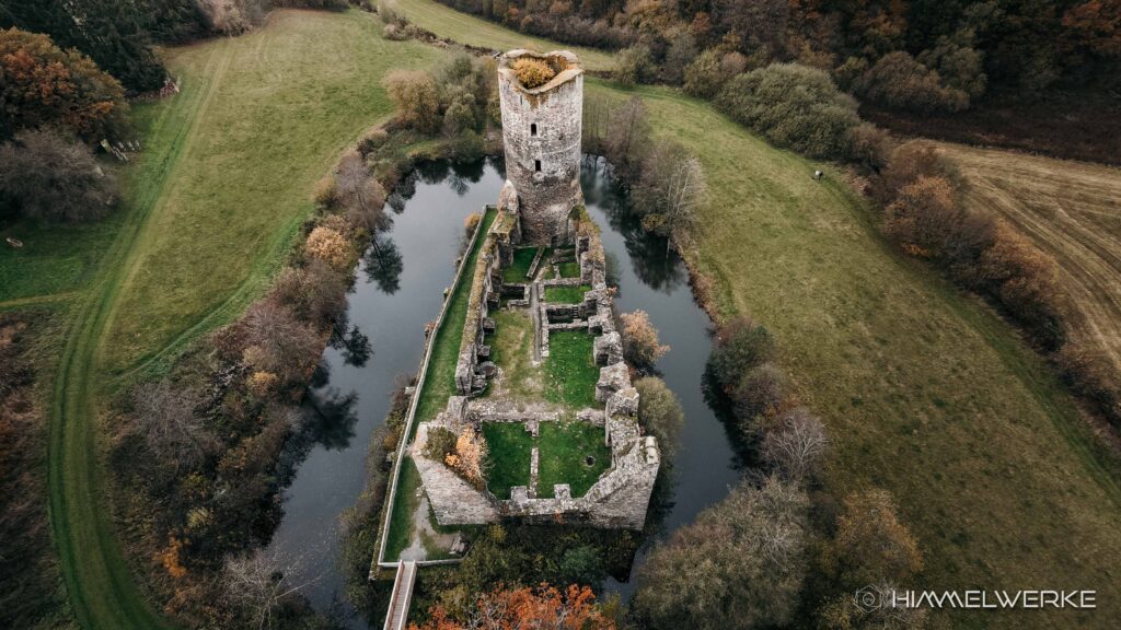 Ein Himmelwerke Bild zum DJI Neo 2 Test. Ein Luftbild, aufgenommen mit der DJI NEO 2. Es ist eine Burgruine zu erkennen und die Kamera schaut schräg nach vorne unten auf die Ruine, sodass man gut ihren Wassergraben und die umliegenden Bäume und Büsche sowie Wiesen erkennen kann. Das bild ist in warme Herbstfarbtöne getaucht.