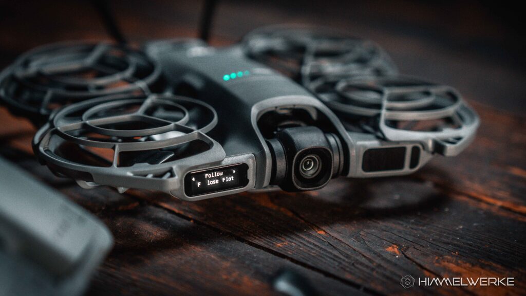 Ein Himmelwerke Bild zum DJI Neo 2 Test. Die DJI Neo 2 auf einerm alten Holzboden von vorne Rechts. Das Display und die BatterieLED leuchten. Es ist der Flugmodus Follow im Display zu erkennen.